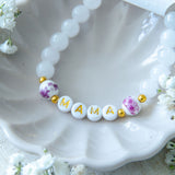 Handmade mama bracelet with white gemstone beads and gold letter beads spelling MAMA, displayed in a ceramic dish with Katie Marie Creations tag and flowers, a thoughtful jewellery gift for mums.