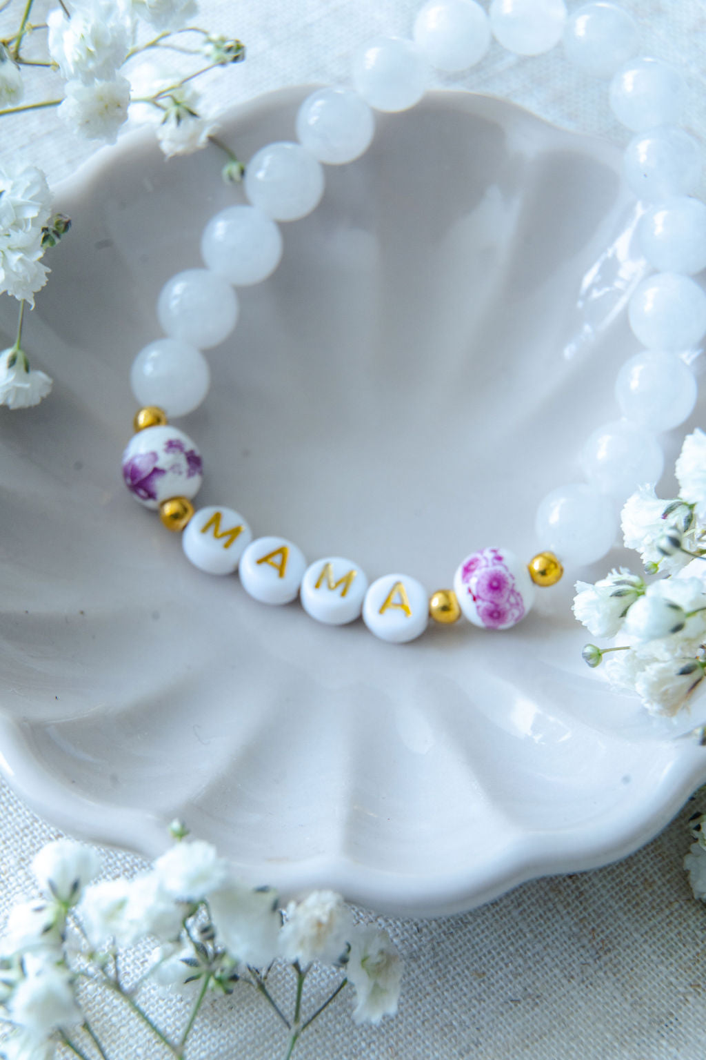 Handmade mama bracelet with white gemstone beads and gold MAMA letter beads, displayed in a ceramic dish with flowers, a thoughtful jewellery gift for new mums or Mother’s Day.