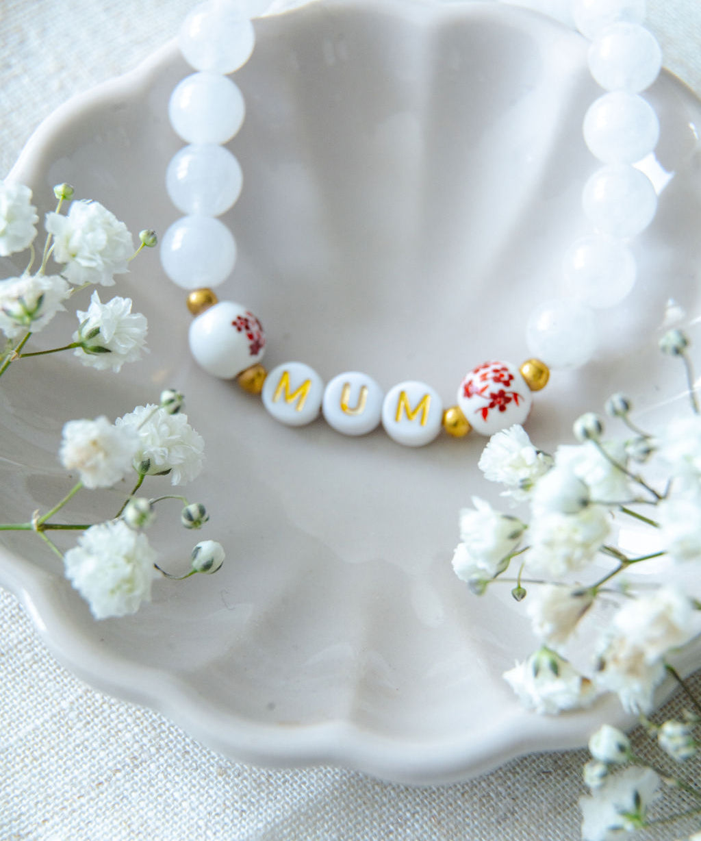 Handmade mum bracelet with white gemstone beads and gold letter beads spelling MUM, styled in a ceramic dish with delicate flowers, a meaningful jewellery gift for mums.