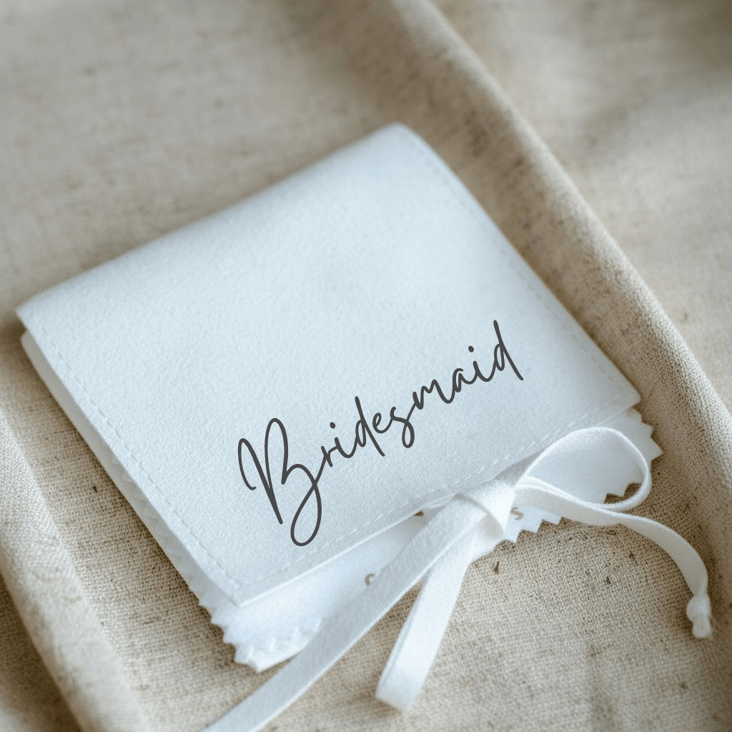 White pouch with 'Bridesmaid' text and a white ribbon on a beige fabric background