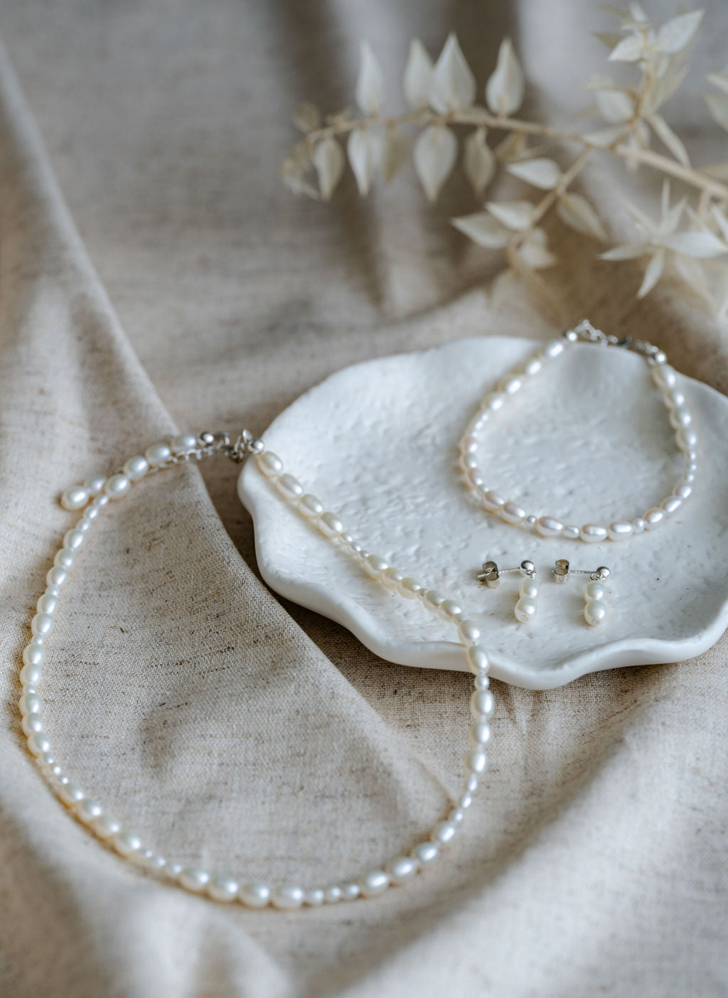 Pearl jewellery set including necklace, bracelet, and earrings on a textured surface with dried leaves.