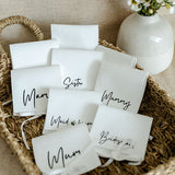 White personalised gift pouches with black text on a woven tray with a vase of flowers in the background