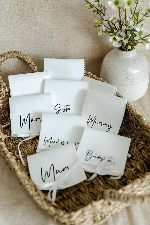 White personalised gift pouches with black text on a woven tray with a vase of flowers in the background
