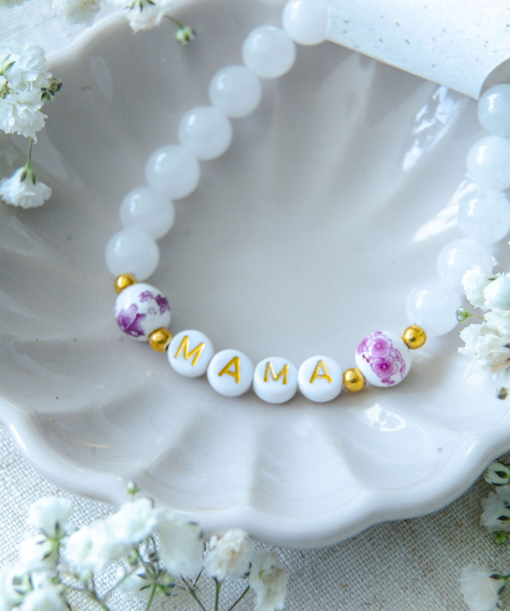 Handmade mama bracelet with white gemstone beads and gold letter beads spelling MAMA, displayed in a ceramic dish with Katie Marie Creations tag and flowers, a thoughtful jewellery gift for mums.