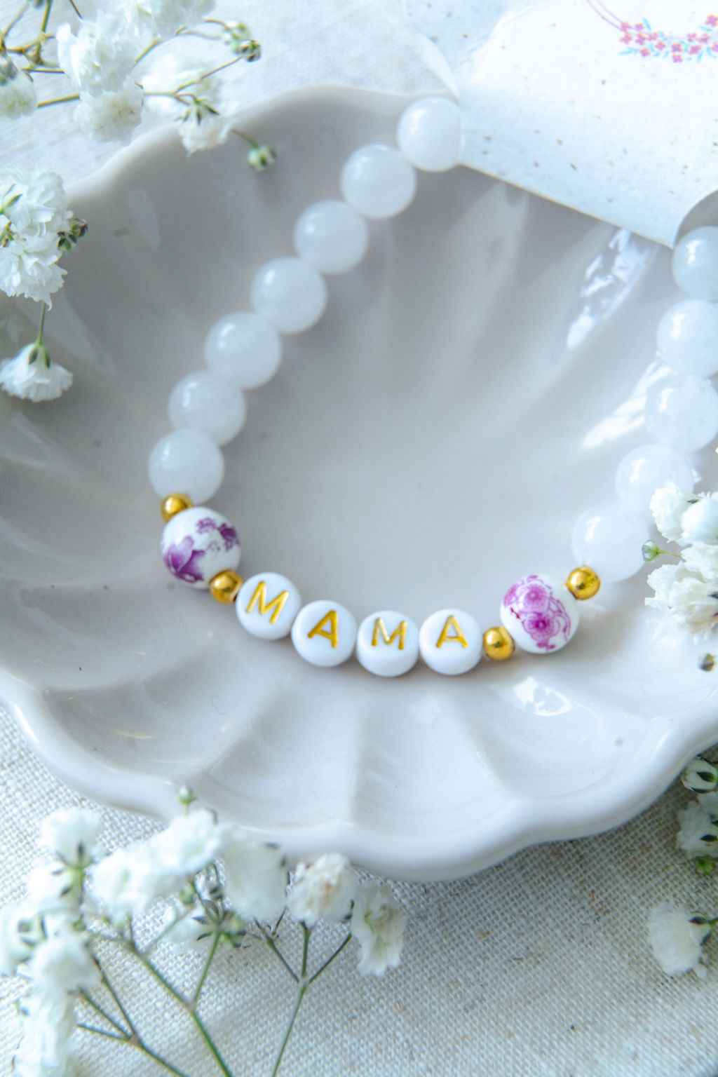 Handmade mama bracelet with white gemstone beads and gold letter beads spelling MAMA, displayed in a ceramic dish with Katie Marie Creations tag and flowers, a thoughtful jewellery gift for mums.