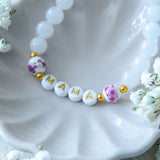 Handmade mama bracelet with white gemstone beads and gold MAMA letter beads, displayed in a ceramic dish with flowers, a thoughtful jewellery gift for new mums or Mother’s Day.