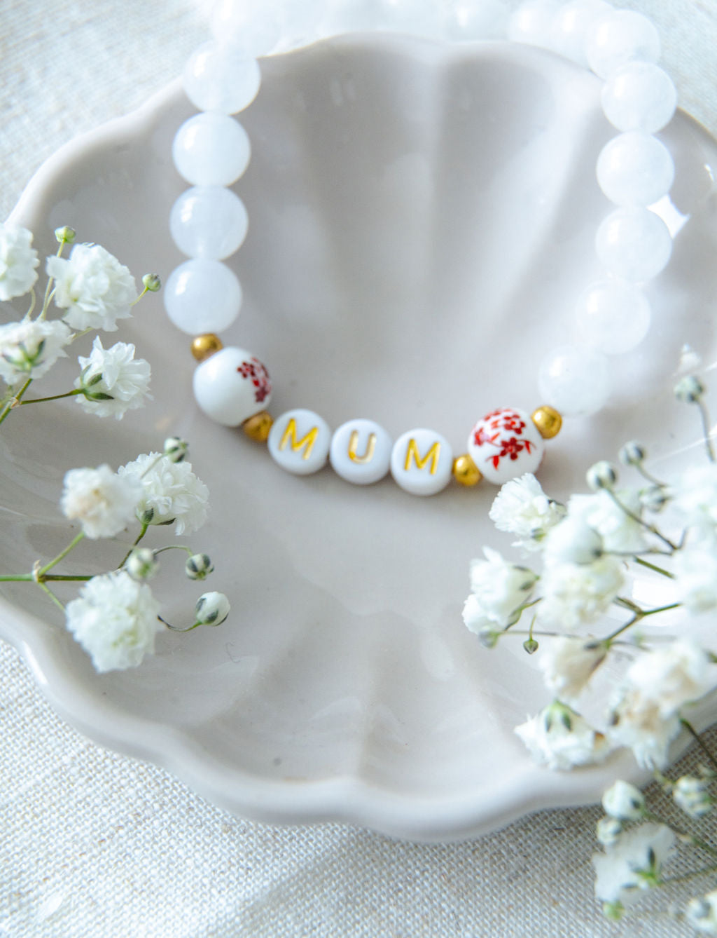 Handmade mum bracelet with white gemstone beads and gold letter beads spelling MUM, styled in a ceramic dish with delicate flowers, a meaningful jewellery gift for mums.