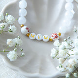Handmade mum bracelet with white gemstone beads and gold letter beads spelling MUM, styled in a ceramic dish with delicate flowers, a meaningful jewellery gift for mums.